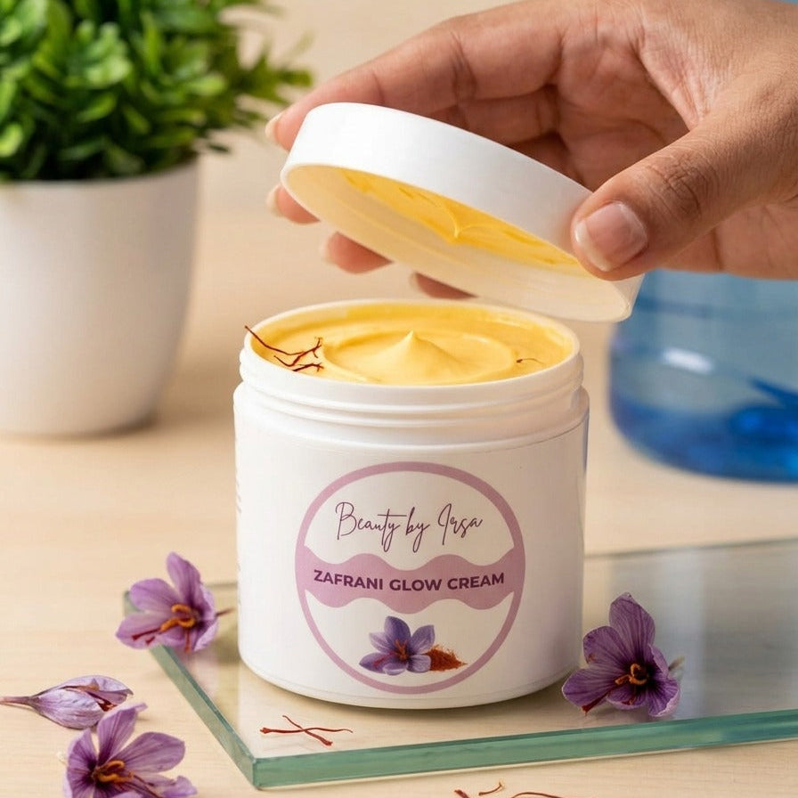 A hand is lifting the lid of a white jar labeled 'Beauty by Rose Zafrani Glow Cream' to reveal yellow cream inside. The jar is placed on a glass tray on a light wooden table, with purple flowers and saffron threads scattered around, a potted plant in the background, and a water bottle blurred on the right.